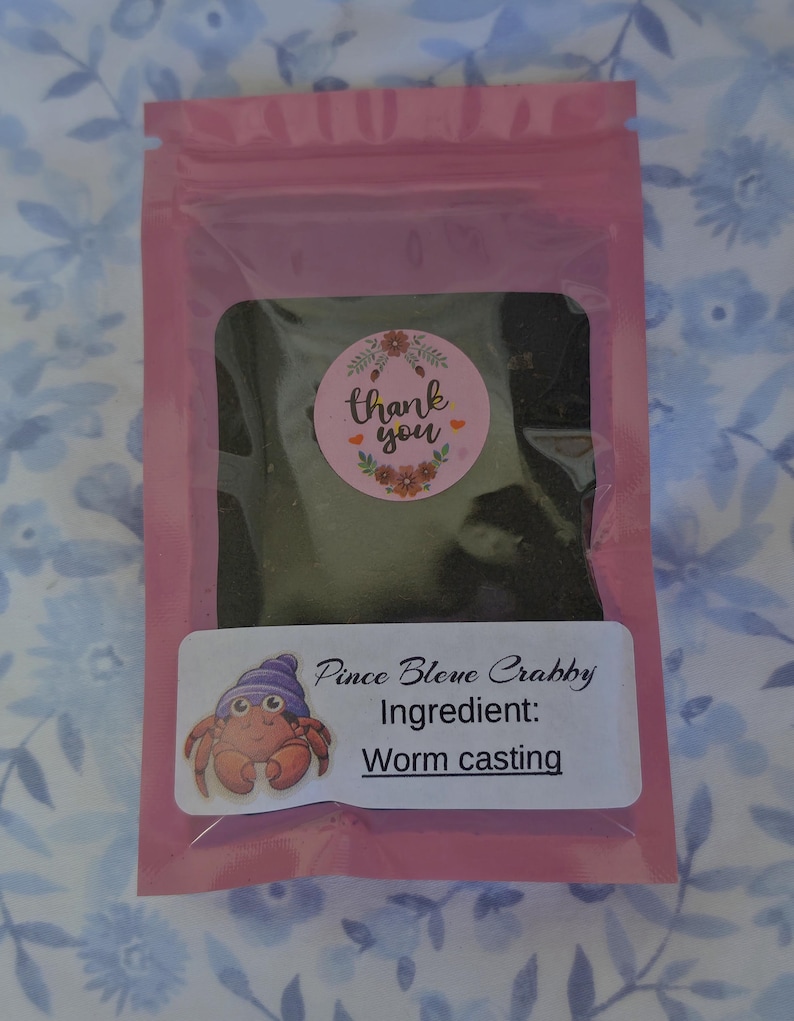 Peut inclure: Un emballage rose contenant une substance sombre, avec un autocollant rond disant "merci" et un motif floral. Une &eacute;tiquette rectangulaire pr&eacute;sente un crabe de dessin anim&eacute; et le texte "Pince Bleve Crabby Ingr&eacute;dient: Worm casting."