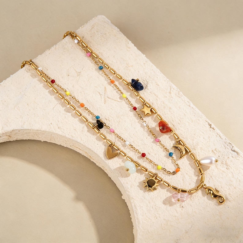 May include: Gold layered necklaces with colourful beads and charms. Charms include a heart, star, sun, moon, seahorse, and a pearl. The necklaces are displayed on a white surface.