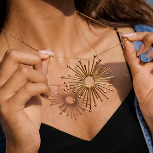 May include: Gold-coloured necklace with a sunburst pendant. The pendant has a central circular design and radiating lines. The necklace hangs from a delicate chain. The pendant's shadow is visible on the black top.