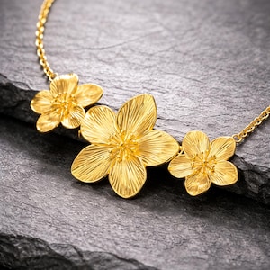 May include: A gold necklace with three flower pendants. Each flower has detailed petals and a central stamen. The necklace is displayed on a dark, textured surface. The gold colour of the necklace is highlighted against the dark background.