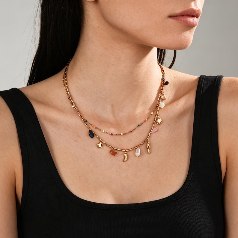May include: A gold-coloured, layered necklace featuring charms such as a star, moon, seahorse, and colourful beads. The necklace is worn against a black top, highlighting the jewellery's design and details.