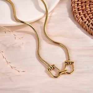 May include: A gold-toned snake chain necklace featuring a central horseshoe-shaped clasp mechanism. The necklace consists of a flexible, rounded metal chain that connects to two symmetrical U-shaped links joined by a central horizontal bar.