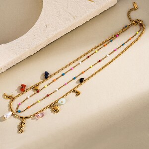 May include: Gold-coloured layered necklaces with colourful beaded accents and various charms. Charms include a seahorse, moon, sun, and other shapes. The necklaces are displayed on a neutral surface, showcasing the jewellery's design and details.