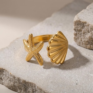 May include: A gold ring with a starfish and seashell design. The ring has an open design, with a starfish on one end and a seashell on the other. The ring is displayed on a light grey stone surface.