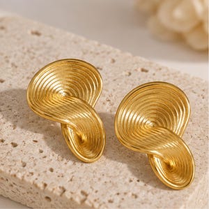 Può includere: Un paio di orecchini color oro caratterizzati da un design a cerchi concentrici a spirale con una superficie scanalata e testurizzata. Gli orecchini presentano una forma asimmetrica e attorcigliata che crea un aspetto stratificato, riflettendo la luce.