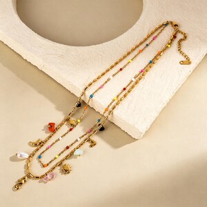 May include: Three gold necklaces with colourful beads and charms. The necklaces feature a variety of charms, including a pearl, a seahorse, a sun, a star, and a heart. The beads are in various colours, including red, orange, yellow, green, blue, and black.