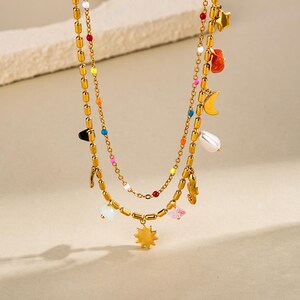 May include: Gold-coloured layered necklace with colourful beads and charms. The necklace features a sunburst pendant, a pearl, a crescent moon, and star charms. The necklace has a delicate chain and is suitable for everyday wear.