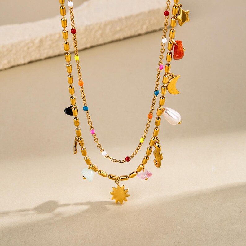 May include: Gold-coloured layered necklace with colourful beads and charms. The necklace features a sunburst pendant, a pearl, a crescent moon, and star charms. The necklace has a delicate chain and is suitable for everyday wear.