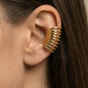 May include: A gold ear cuff with a spiral design. The ear cuff wraps around the ear, creating a bold and modern look. The jewelry is made of a shiny, gold-colored metal.