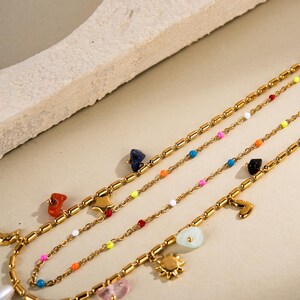 May include: Several gold necklaces with colourful beads and charms are displayed. The necklaces have charms such as a star, sun, and heart. The beads are in various colours, including blue, orange, pink, and yellow.