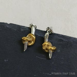 Op de afbeelding: Een paar oorknopjes met een tweekleurig ontwerp, voorzien van een gepolijste zilverkleurige stift en een goudkleurig knoopelement. De oorbellen hebben een gladde, reflecterende metalen afwerking en zijn gelabeld als Modern Knot Studs.
