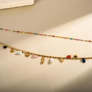 May include: Two gold necklaces. The top necklace has small, colourful beads. The bottom necklace has charms including a heart, sun, seahorse, and a small car. The charms are in various colours.