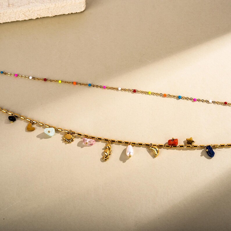 May include: Two gold necklaces. The top necklace has small, colourful beads. The bottom necklace has charms including a heart, sun, seahorse, and a small car. The charms are in various colours.
