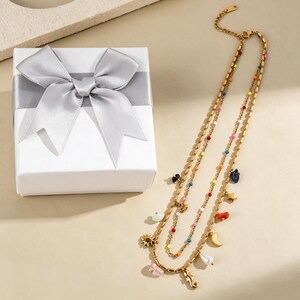 May include: A gold-tone, multi-strand necklace featuring colourful beads and charms, such as a seahorse, sun, and moon. It is presented beside a white gift box tied with a silver ribbon.