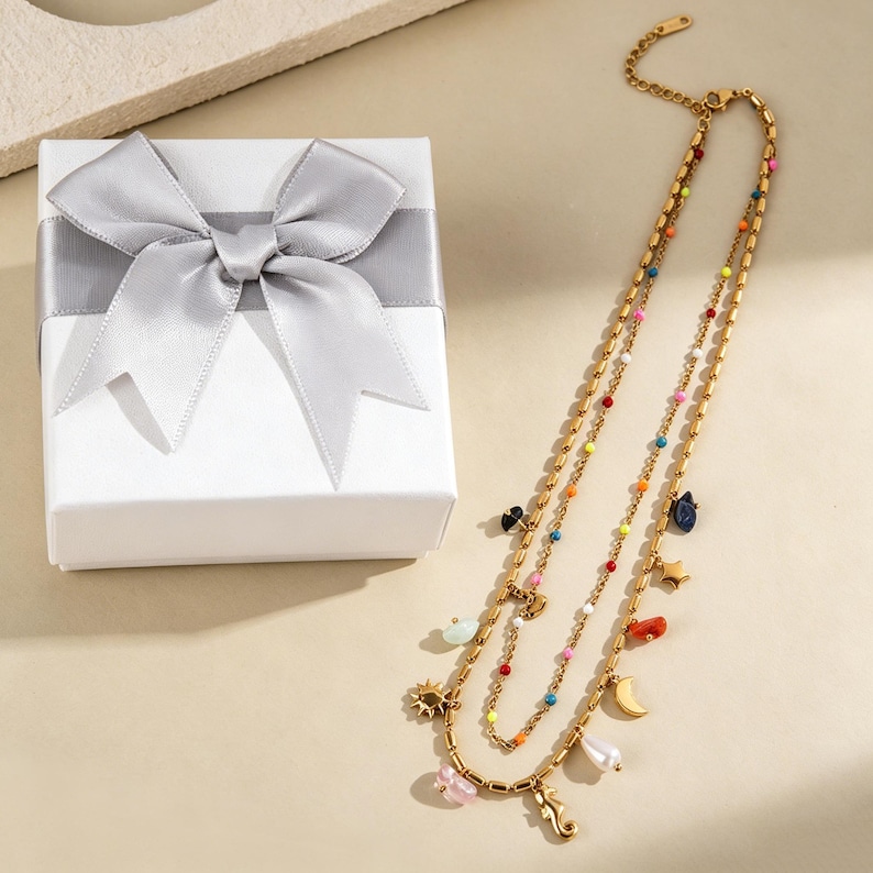 May include: A gold-tone, multi-strand necklace featuring colourful beads and charms, such as a seahorse, sun, and moon. It is presented beside a white gift box tied with a silver ribbon.