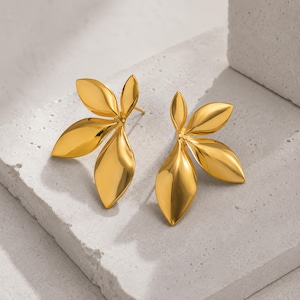 May include: A pair of gold-toned earrings shaped like clusters of three smooth, polished leaves. Each earring features a central stem connecting three distinct, rounded leaf forms with a reflective, metallic finish and curved edges.