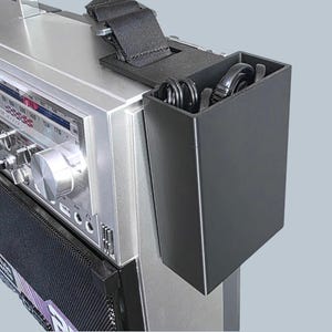 May include: A black rectangular storage container attached to the side of a silver boombox with a black fabric strap. The container holds coiled black cables. The boombox features a silver control panel with a tuning dial and various ports.
