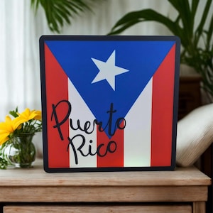 May include: Square sign featuring the Puerto Rican flag design with a white star on a blue triangle, red stripes, and the word "Puerto Rico" in script. The sign has a black border and is displayed on a wooden surface.