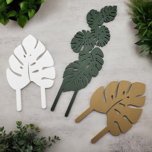 Monstera trellis plant stakes in two styles (single leaf and six-leaf tower) and three colors (white, green, and tan) with plants around the edges.