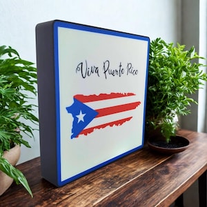 May include: A rectangular sign with a white background, blue border, and dark gray sides. The sign features the text "Viva Puerto Rico" in cursive above a stylized Puerto Rican flag.