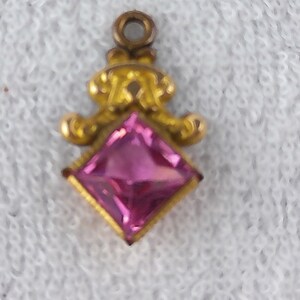 May include: A small, gold-colored pendant with a square, pink gemstone at its center. The pendant has an ornate design above the gemstone, with a small loop at the top for attaching to a chain or bracelet. The gemstone is a vibrant pink color.
