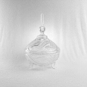 May include: A clear glass candy dish with a lid and three small feet. The dish has a decorative, swirling pattern on the body and lid. The lid has a small, vertical handle. The dish is displayed against a white background.