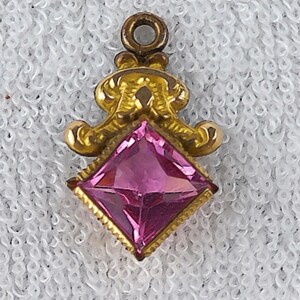 May include: A gold-toned pendant with a pink, square-cut gemstone. The pendant has an ornate, filigree design surrounding the gem and a small loop at the top for a chain. The gemstone is a vibrant pink color.
