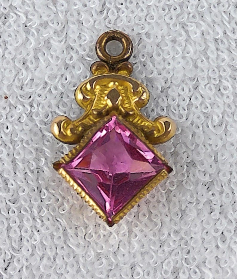 May include: A gold-toned pendant with a pink, square-cut gemstone. The pendant has an ornate, filigree design surrounding the gem and a small loop at the top for a chain. The gemstone is a vibrant pink color.