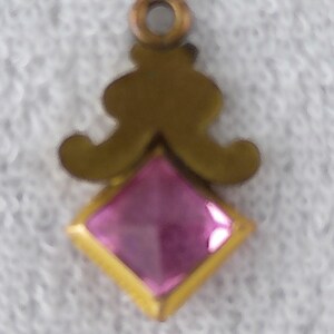 May include: A small, ornate pendant with a gold-colored metal frame. The pendant features a square, faceted pink gemstone set within a gold border. Above the gemstone is a decorative gold design, and a small loop for hanging.