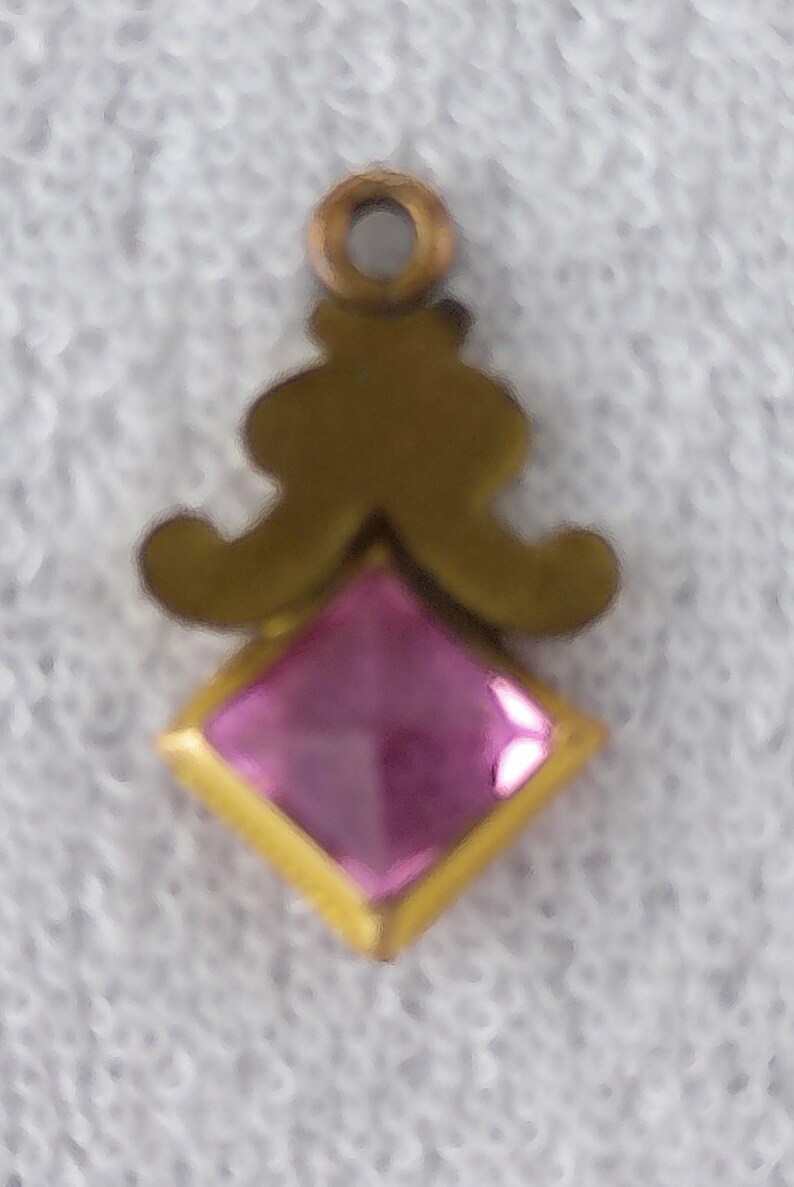May include: A small, ornate pendant with a gold-colored metal frame. The pendant features a square, faceted pink gemstone set within a gold border. Above the gemstone is a decorative gold design, and a small loop for hanging.