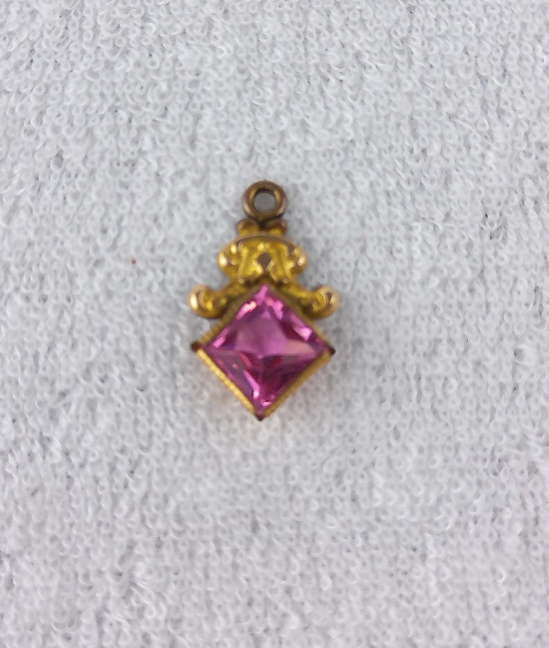 May include: A small, gold-colored pendant with a square, pink gemstone at its center. The pendant has an ornate design above the gemstone, with a small loop at the top for attaching to a chain or bracelet. The gemstone is a vibrant pink color.