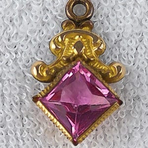 May include: A gold-toned pendant with a pink, square-cut gemstone. The pendant has an ornate, scrolled design above the gem and a small loop at the top for a chain. The gemstone is a vibrant pink color.