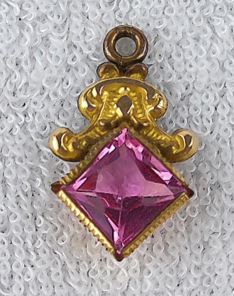 May include: A gold-toned pendant with a pink, square-cut gemstone. The pendant has an ornate, scrolled design above the gem and a small loop at the top for a chain. The gemstone is a vibrant pink color.