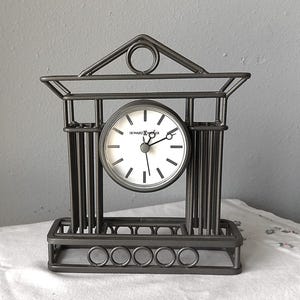 Pode incluir: Um relógio de metal preto com um mostrador branco e ponteiros pretos. O relógio tem a forma de um edifício com um topo decorativo. O mostrador do relógio diz "Howard Miller".