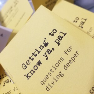 May include: A stack of yellow cards with the text "Getting' to know ya, pal" and "questions for diving deeper" printed on them. The cards are part of a game or activity designed to help people get to know each other better.