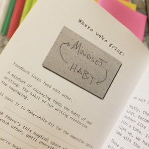 May include: A close-up of a page from a book with the text "Where we're going: Mindset Habit" written in black marker on a gray sticky note. The text on the page reads "Feedback loops feed each other. A mindset of naysaying feeds the habit of not writing. The habit of not writing reinforces the naysaying."
