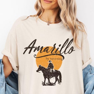 May include: A beige t-shirt with the text "Amarillo by morning" in black with a vintage style font. The text is above a silhouette of a cowboy riding a horse in front of a yellow sun.