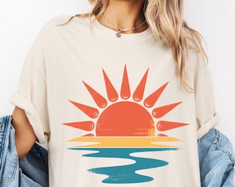 May include: Ivory-colored t-shirt featuring a sunset design. The graphic depicts a vibrant orange sun with yellow rays above a blue and yellow horizon line. The word "IVORY" is printed at the bottom left of the image.