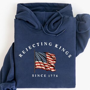 May include: Navy blue hoodie with the text "Rejecting Kings Since 1776" and an American flag graphic. The hoodie has a drawstring hood and a kangaroo pocket. The word "Navy" is displayed in a starburst graphic.