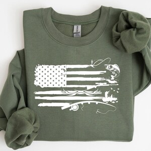 May include: Military green sweatshirt featuring a distressed white graphic of the American flag with fishing-related imagery. The design includes stars, stripes, fish, and fishing rods. The sweatshirt has a crew neck and long sleeves.
