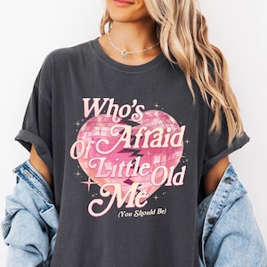 May include: Dark gray t-shirt with the text "Who's Afraid of Little Old Me (You Should Be)" in pink and white. The graphic is a heart-shaped disco ball. The shirt has short sleeves and a relaxed fit.