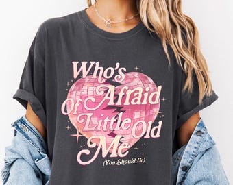 May include: Dark gray t-shirt with the text "Who's Afraid of Little Old Me (You Should Be)" in pink and white. The graphic is a heart-shaped disco ball. The shirt has short sleeves and a relaxed fit.