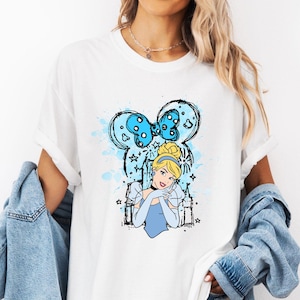 May include: White t-shirt with a blue and white illustration of Cinderella with a bow on her head and a castle in the background. The illustration is surrounded by stars and hearts.