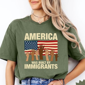 May include: A green t-shirt with the text "America was built by immigrants" and an American flag with raised hands of different skin tones.