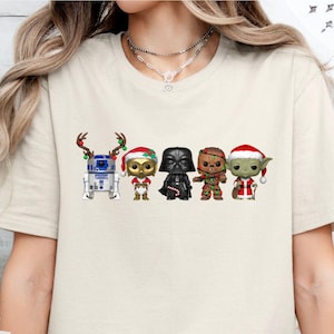May include: A sand-colored t-shirt featuring a graphic print of five stylized characters in holiday attire. The characters include a robot with reindeer antlers, a small creature in a Santa hat, a black helmeted figure, a brown creature, and a green alien.