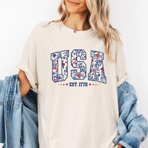 May include: Ivory-colored t-shirt with the word "USA" in a floral design, blue outline, and the text "EST. 1776" below. The shirt is paired with a light blue denim jacket.