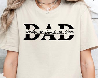May include: Sand-colored t-shirt with the word "DAD" in bold black letters. Below the word "DAD" are the names "Emily Sarah Jane" in a smaller font, separated by heart symbols. The shirt is a crew neck.