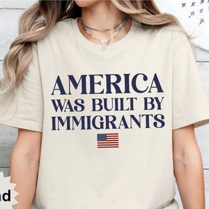 May include: A beige t-shirt with the text "America was built by immigrants" in navy blue lettering. A small American flag is printed below the text.