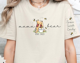 May include: A beige t-shirt with a Winnie the Pooh illustration and the text "mama bear est. 2025". The shirt also has the names "Grace, Sarah, Emily" written on the right sleeve.