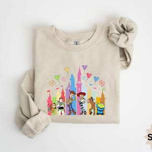 Peut inclure: Sweat-shirt couleur sable avec un graphisme color&eacute; des personnages de Toy Story devant un ch&acirc;teau avec des feux d'artifice et des ballons. Le sweat-shirt a des manches longues et un col rond. Le mot "Sand" est imprim&eacute; en bas &agrave; droite.